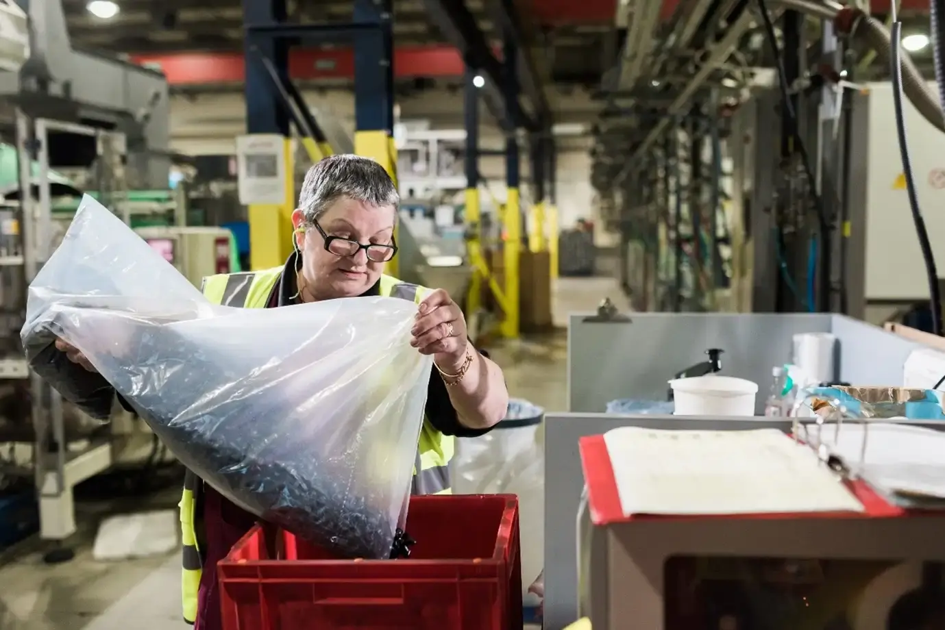 A worker in a safety vest handles a large plastic bag of materials in an industrial setting, surrounded by machinery and equipment.