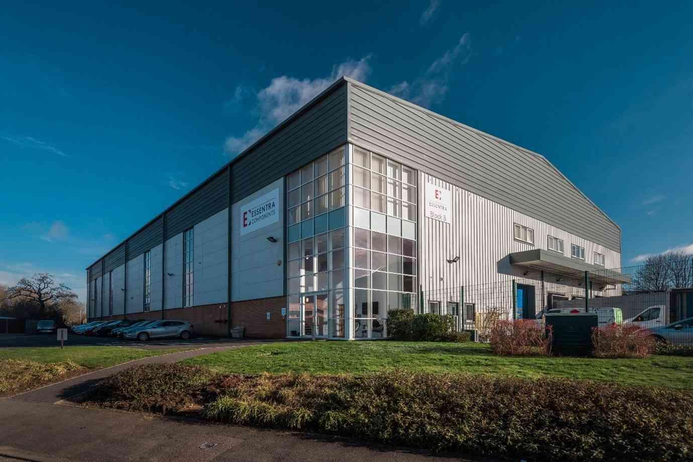 Large industrial warehouse with metal siding, glass entry, and a loading dock. Surrounded by grass and parked cars under a clear blue sky.