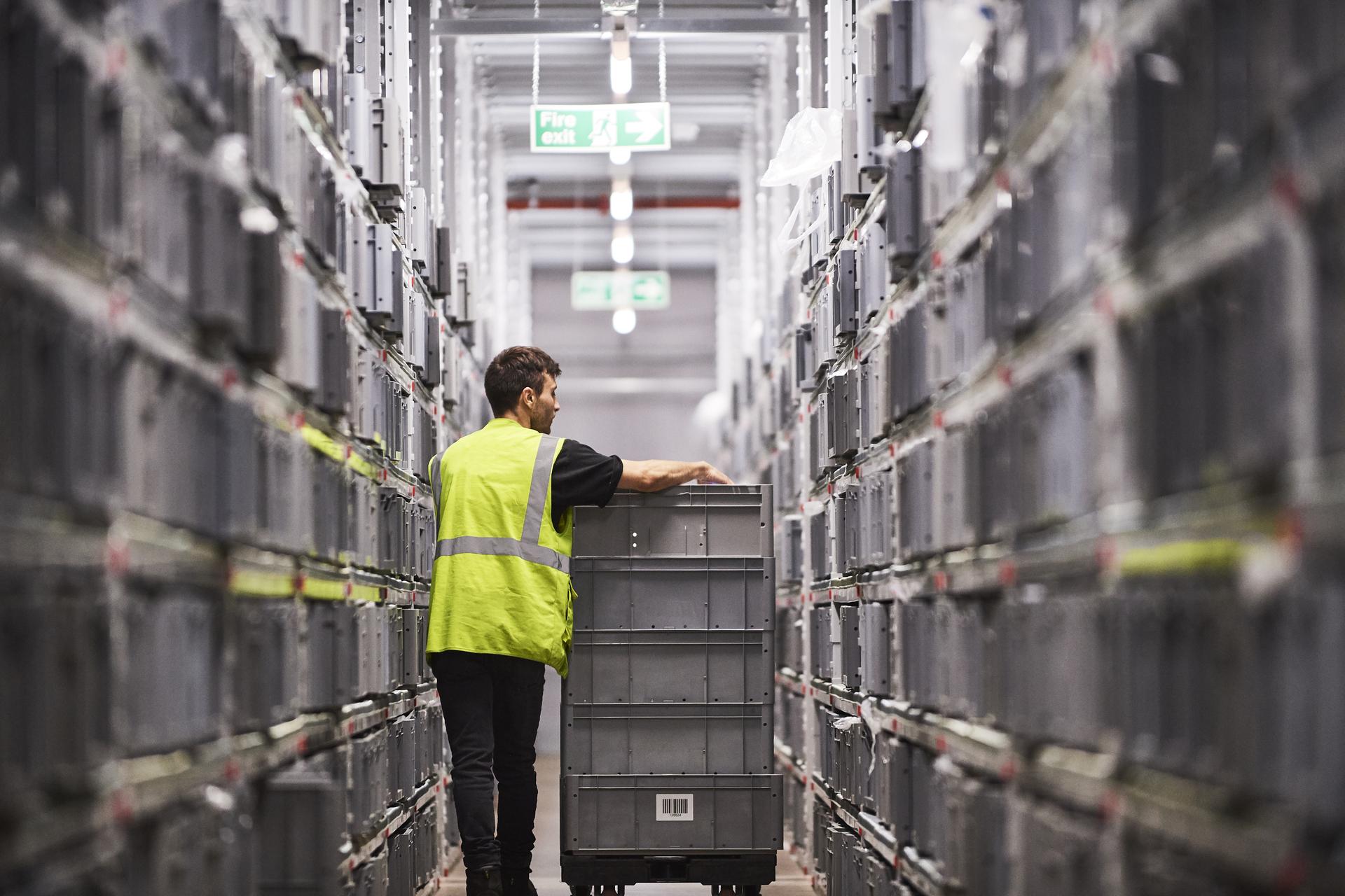 Worker in a yellow vest moves a cart through a warehouse aisle lined with stacked bins under bright lighting.