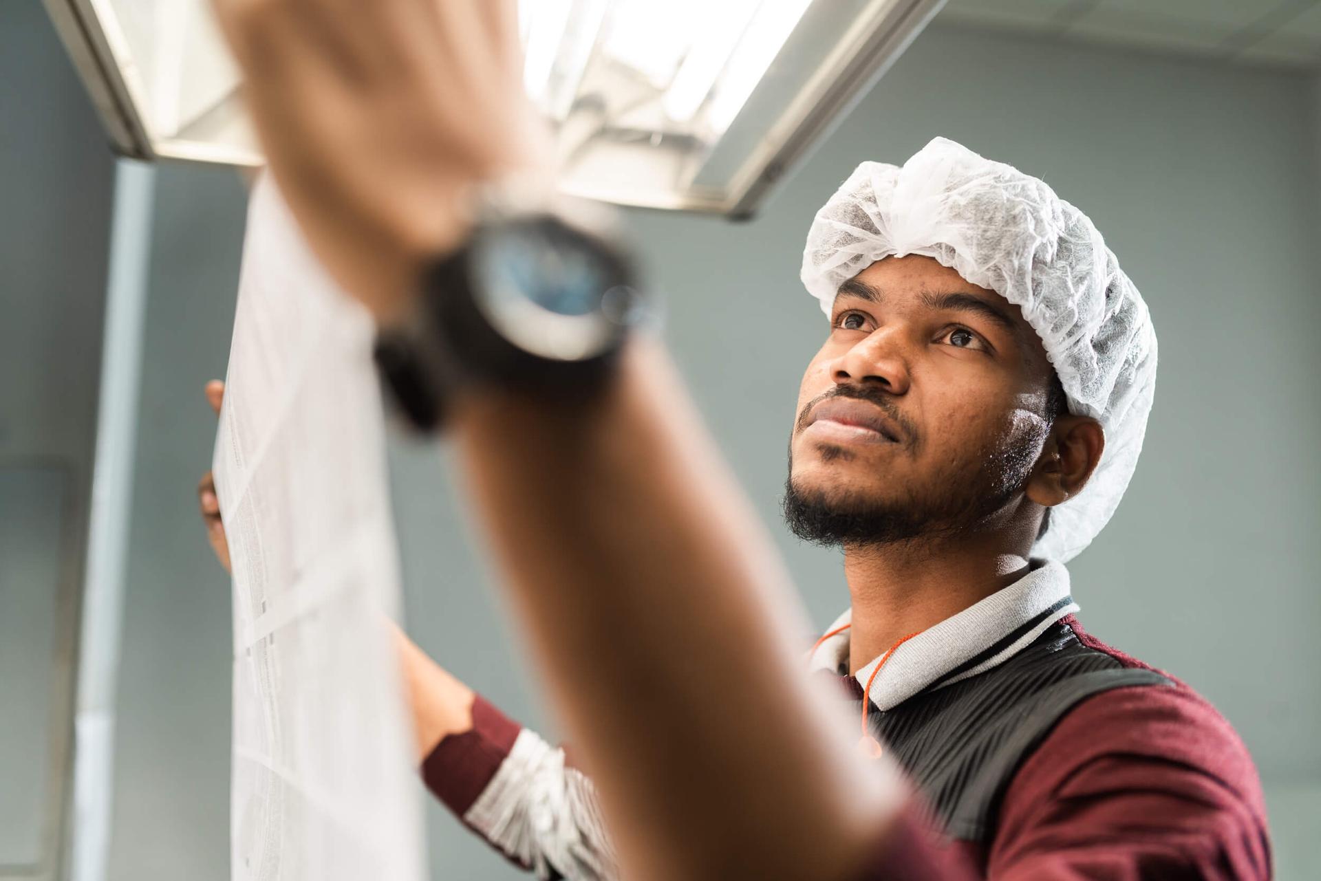 A man wearing a hairnet and a wristwatch inspects a translucent sheet under a bright light, in a focused industrial setting.